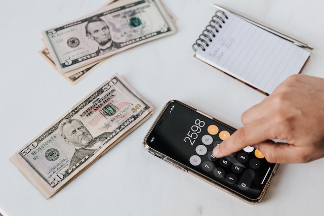 Two stacks of American money on a table next to a small notepad and an iPhone open to the calculator.